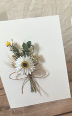Floral corsage with a white flower and yellow center on a white card with a wooden surface background.