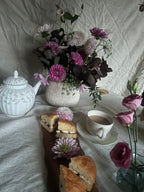 A styled still life scene on a white fabric backdrop featuring a white floral teapot, a woven vase filled with pink, purple, and dark foliage flowers, a cup of tea on a saucer, and sliced bread on a wooden board with a flower placed in front.